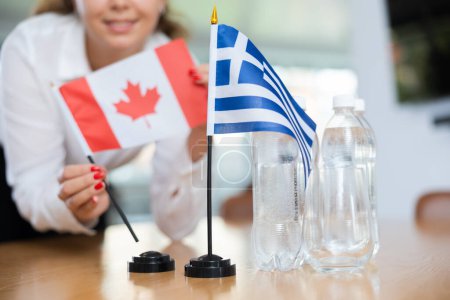 Unerkennbare unkonzentrierte Frau, die Raum für internationale Verhandlungen und Kommunikationsdiskussionen von Führungspersönlichkeiten schafft. Lady stellt Miniaturen von Flaggen Griechenlands und Kanadas auf den Tisch.