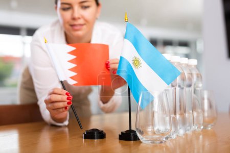 Little flag of Argentina on table with bottles of water and flag of Bahrain put next to it by positive young woman 