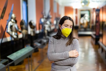 Interested young woman wearing yellow protective face mask viewing collection of medieval knights armor in historical museum. Forced precautions in pandemic