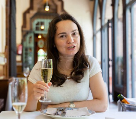 Portrait of slightly drunk laughing woman sitting at table in restaurant with glass of sparkling wine