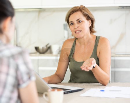 Dos mujeres de mediana edad positivas teniendo conversación de negocios mientras están sentadas en la mesa de la cocina