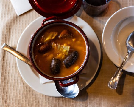 There is traditional Spanish dish on table - Fabada Asturian. Stewing soup cooking of beans, sausages and spices in clay pot