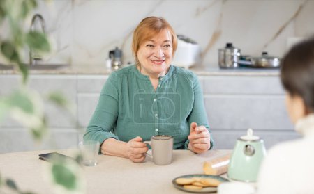 Daheim in der Küche am Tisch sitzt eine ältere lächelnde Frau und hat eine interaktive Beziehung zu einem Freund und trinkt Kaffee oder Tee. Konzept lebendige Beziehung und persönliche Gespräche