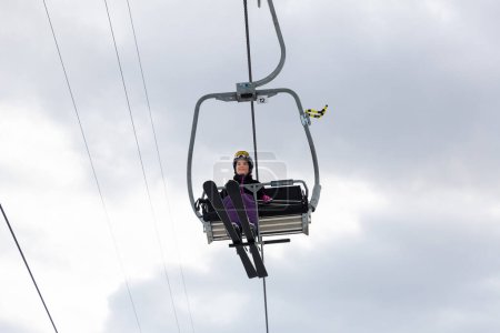 Smiling young woman in ski outfit sitting alone on chairlift, raising to slopes for skiing on background of cloudy winter sky, bottom view