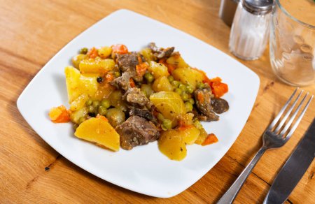 There is portion of stewed veal with vegetables in deep plate. Large pieces of potatoes and carrots soaked in gravy and spices appetizingly complement tender meat