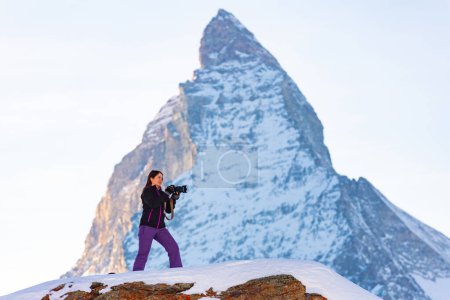 Mujer interesada fotógrafa de pie sobre roca cubierta de nieve con pico de Matterhorn en el fondo capturando vistas panorámicas de la montaña de los Alpes suizos en el día de invierno