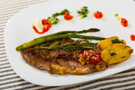 Roasted veal steak served with baked vegetables, adjika and garlic herbs sauce on white plate on striped background..