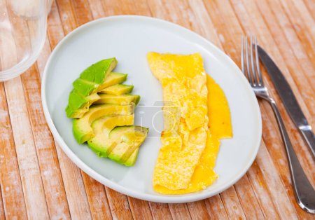 La idea para un desayuno saludable y bajo en carbohidratos es tortilla y aguacate medio picado. Plato simple se sirve con electrodomésticos y vaso de aperitivo.