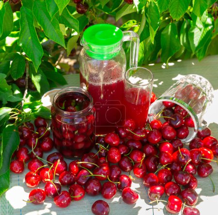 Closeup of fresh ripe red sweet cherries, mason jar of compote with berries and carafe with cherry juice outdoors in shade of green foliage of fruit trees in orchard..