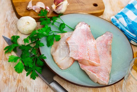 Raw fish with condiments, red perch fillet on plate with garlic and parsley on wooden surface