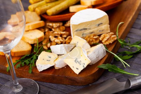On cutting board there is slicing of blue cheese and rusk, complemented with some walnuts and decorated with fresh arugula leaves. Large platter with appetizer stands on wooden table next to glasses