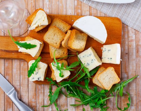 En la tabla de cortar hay rebanadas de queso azul y rusk, complementadas con hojas de rúcula frescas. Tablero grande con aperitivo sobre mesa de madera junto al vidrio