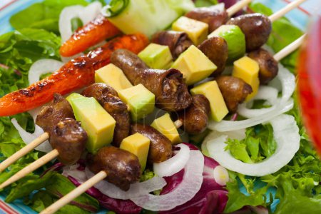 Closeup of grilled chicken hearts on skewers with avocado, baked carrots and fresh greens on colored plate