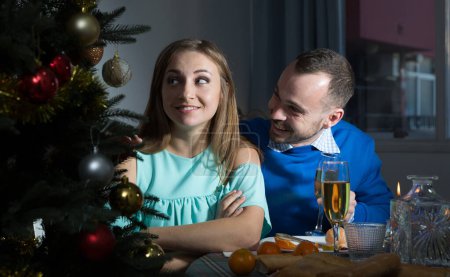 Ingratiating smiling man apologizing to girlfriend, pretending that she is offended while sitting at home table at Christmas night..