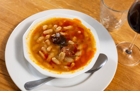 There is traditional Spanish dish on table - Fabada Asturian. Stewing soup stew of beans, sausages and spices in plate