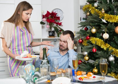 Joven mujer disgustada limpiando mesa después de la celebración de Navidad y regañando hombre borracho sentado a la mesa