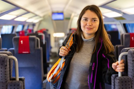 Smiling female backpacker traveling by train, standing in aisle between rows of seats holding on handle ..