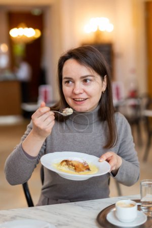 Cheerful woman enjoying traditional apple strudel with vanilla sauce and fragrant espresso in coffee house while traveling in Vienna