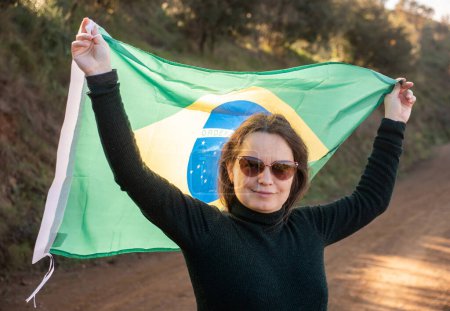 Smiling middle-aged woman holding large flag of Brazil against the background of ground road