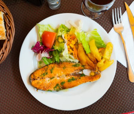 Es gibt große Scheiben Lachs auf dem Grill auf dem Teller gekocht. Fisch wird mit einer Portion Pommes frites und Salat mit frischem Gemüse ergänzt.