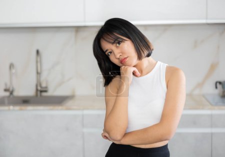 Decepcionado joven asiática dama de pie por sillas en la cocina con armarios blancos limpios