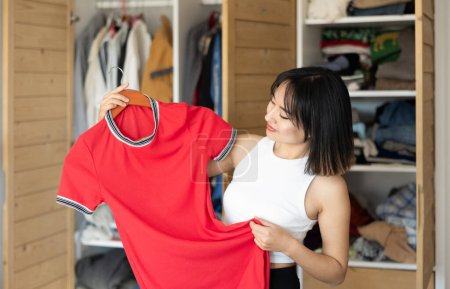 Zufriedene junge Asiatin, die vor Kleiderschrank steht und entscheidet, welche Kleidung sie bevorzugt