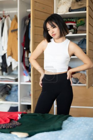 Distempered joven asiática dama de pie delante de armario decidir qué ropa a preferir