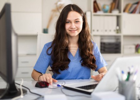 Una joven doctora en prácticas que está pasando por una pasantía en una clínica médica está sentada en el lugar de trabajo en una computadora