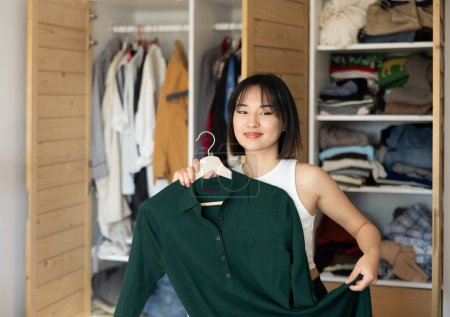 Frohe junge Asiatin, die Kleider in der Hand hält, um in der Umkleidekabine die Wahl zu treffen