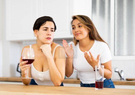 Joven latina consolando a su hermana molesta mientras está de pie en la cocina casera con vasos de vino y discutiendo problemas