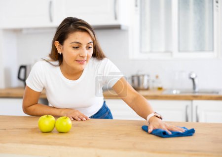 Retrato de alegre joven latina limpiando mesa con paño y spray en la cocina. Concepto de tareas domésticas cotidianas