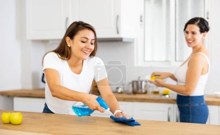 Smiling young Hispanic woman wiping dust from kitchen table surfaces with cleaning rag and detergent spray while cleaning house with her girlfriend
