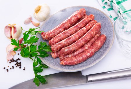 Raw spicy longaniza sausages from minced pork prepared for frying lying on plate with knife, garlic, peppercorns and fresh parsley. Traditional Spanish specialty