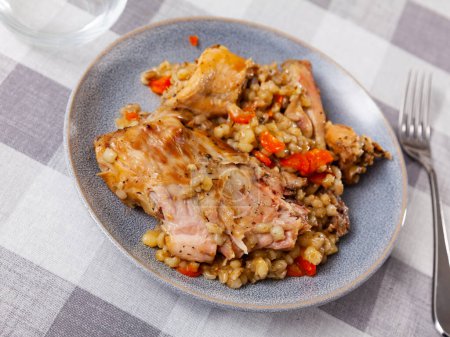 Portion of hearty lunch on plate - pieces of baked rabbit competed with pearl barley and carrot.