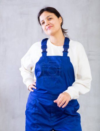 Positive young woman, professional construction worker, dressed in blue jumpsuit posing confidently against gray studio background, looking at camera with smile