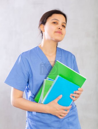 Positive confident young woman, professional doctor, in blue uniform holding medical records. Studio photoshoot on gray background