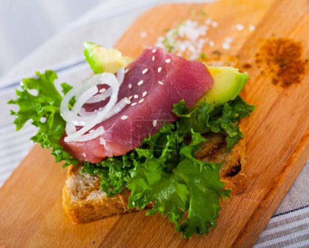 Tostadas saludables con atún crudo y aguacate maduro adornado con verduras frescas en tabla de cortar de madera