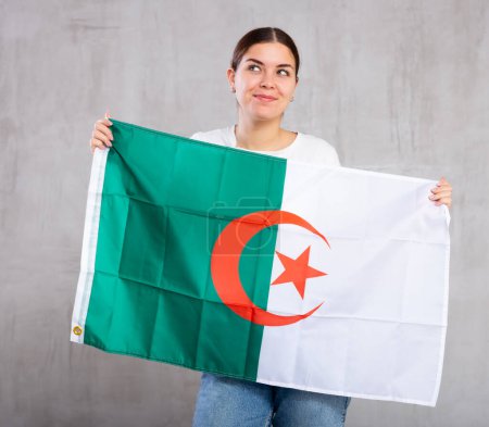 Joyeuse jeune femme avec drapeau algérien dans les mains posant joyeusement sur fond unicolore clair