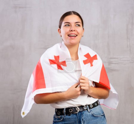 Joyous young woman with Georgia flag on shoulders posing happily against light unicoloured background