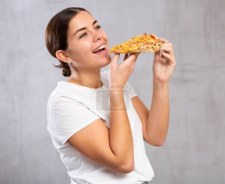 Photo of pleased young woman biting a piece of pizza held in hands against light unicoloured background