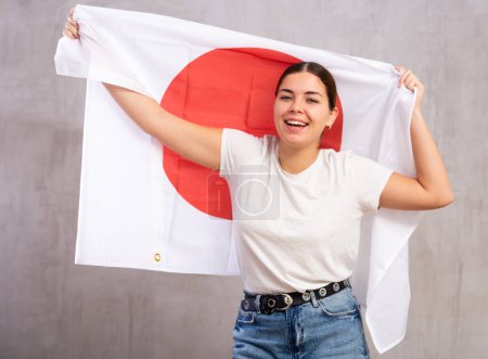 Joyful girl stands with flag of Japan in her hands. Isolated on gray background