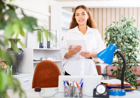 Cheerful young business woman standing in office with papers in hands. Concept of favourable business offer, profitable proposition or great deal
