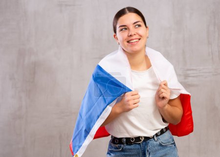 Joyous young woman with Czech flag on shoulders posing happily against light unicoloured background