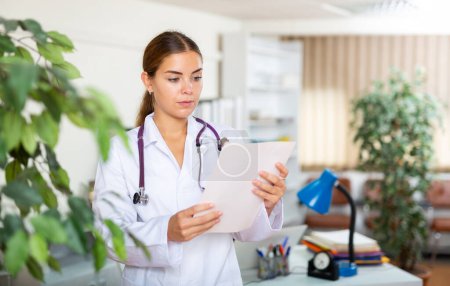 Jeune femme médecin en robe blanche et stéthoscope sur les épaules debout dans le hall de la clinique. Shes tenant le document entre les mains.