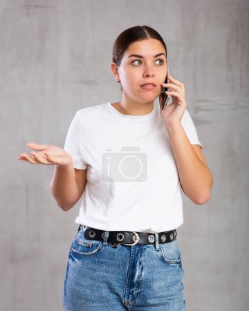 Foto einer erstaunten jungen Frau, die mit Sorge vor grauem, schattenlosen Hintergrund auf dem Handy spricht