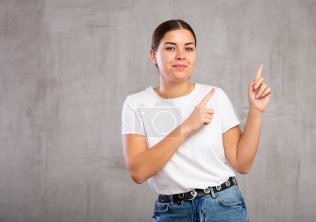 Photo of cheery young woman pointing fingers away against gray shadeless background