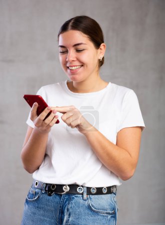 Porträt einer fröhlichen jungen Frau, die vor hellem, einfarbigem Hintergrund freudig aufs Handy blickt