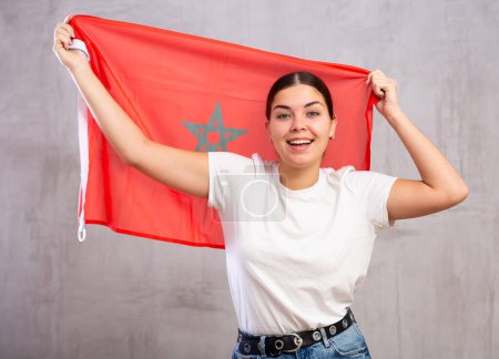 Happy young woman in casual clothes posing and smiling, standing with flag of Morocco against gray wall