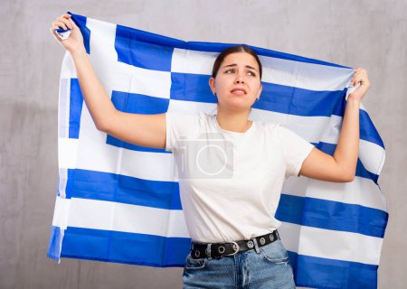 Sad young woman with Greece flag in hands posing sorrowfully against light unicoloured background