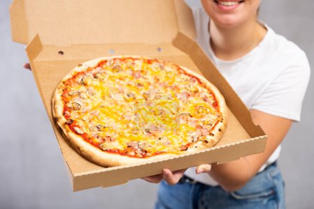Lächelnde junge Frau hält vor hellem einfarbigem Hintergrund ganze Pizza in Schachtel